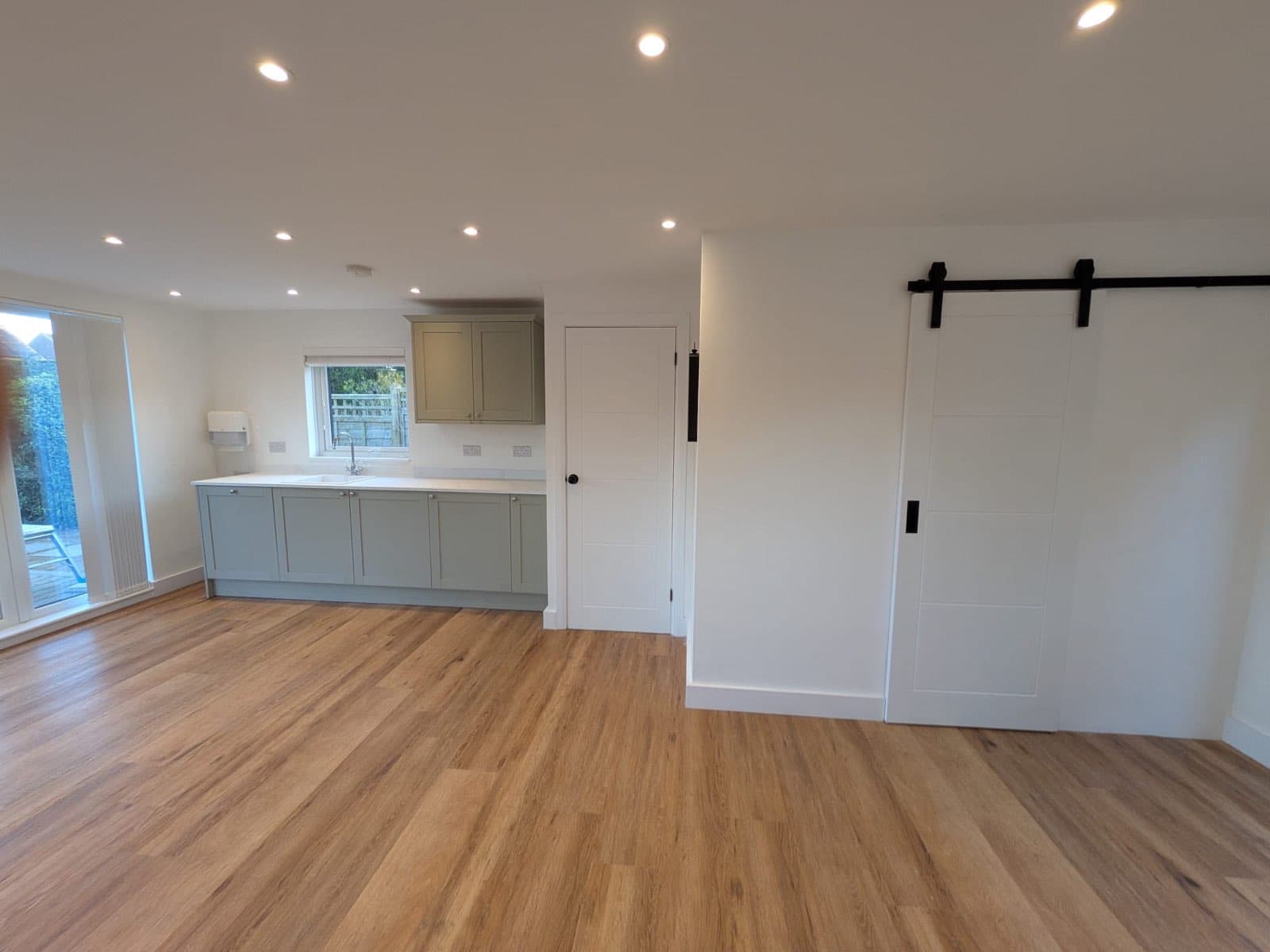 Inside a large open-plan garden room with a sage green kitchen, white barn-style sliding door, oak-effect vinyl floor and recessed downlights