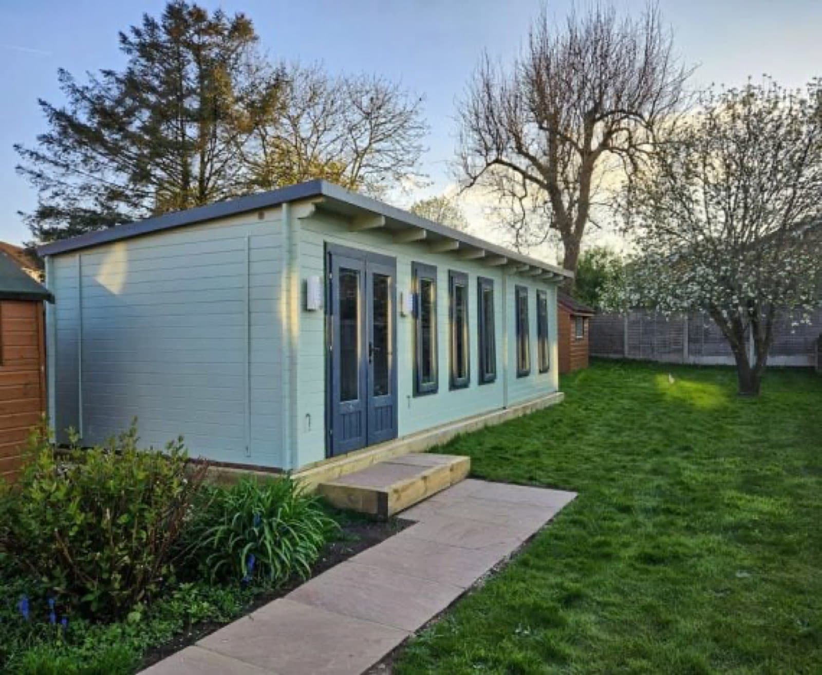 Pale blue painted pent log cabin with glazed double doors and a long run of windows, on a lawn with a paved path up to it