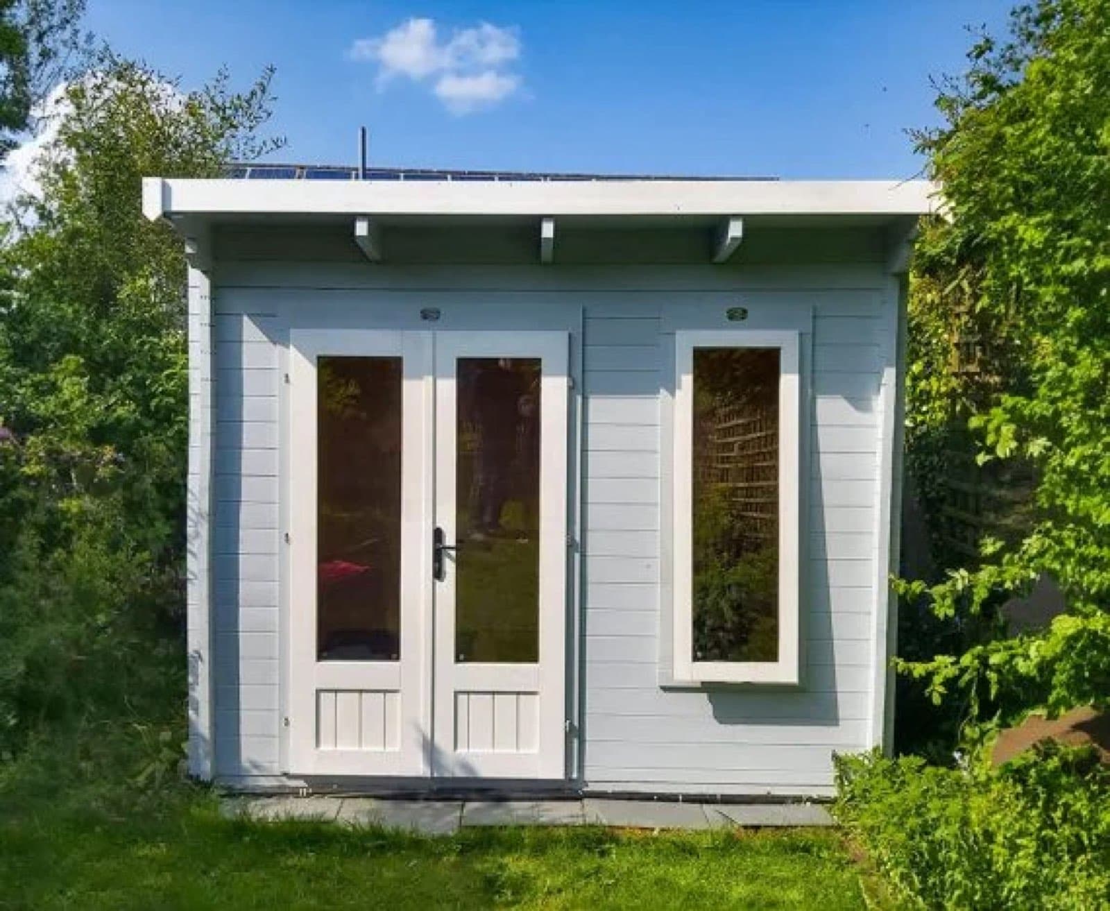 Pale blue and white painted pent cabin with glazed French doors, surrounded by greenery