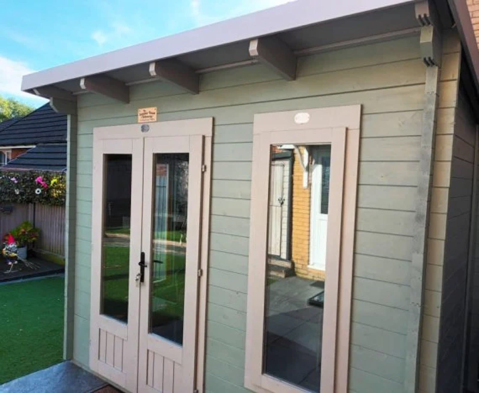 Sage green painted log cabin with cream doors and window surrounds, beside artificial lawn