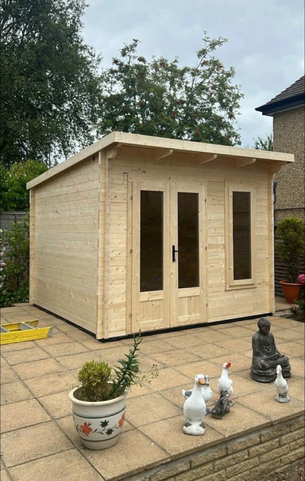 Untreated pent log cabin with glazed double doors and a side window, on a paved patio with planters and a stone Buddha