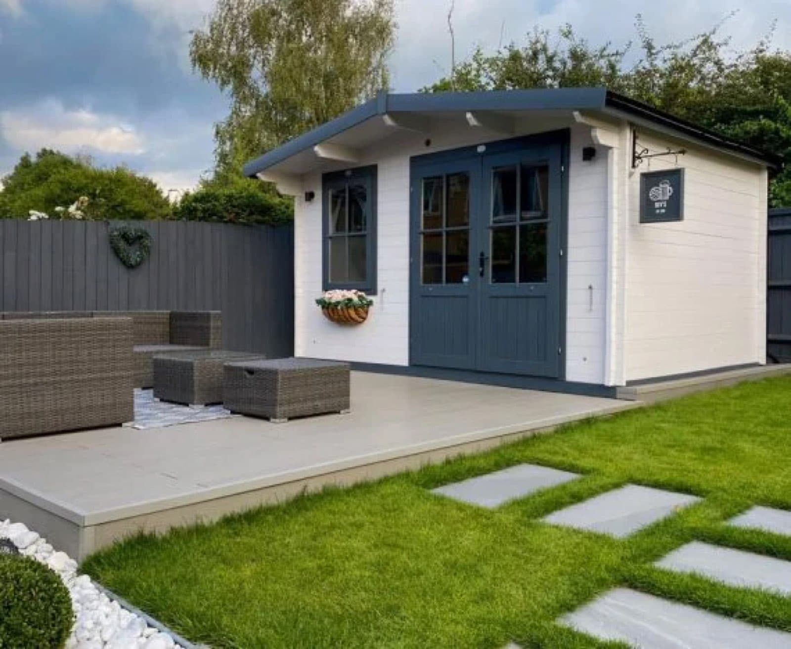 White painted log cabin with navy doors and an apex roof, on a porcelain patio with a rattan sofa and stepping stones across the lawn