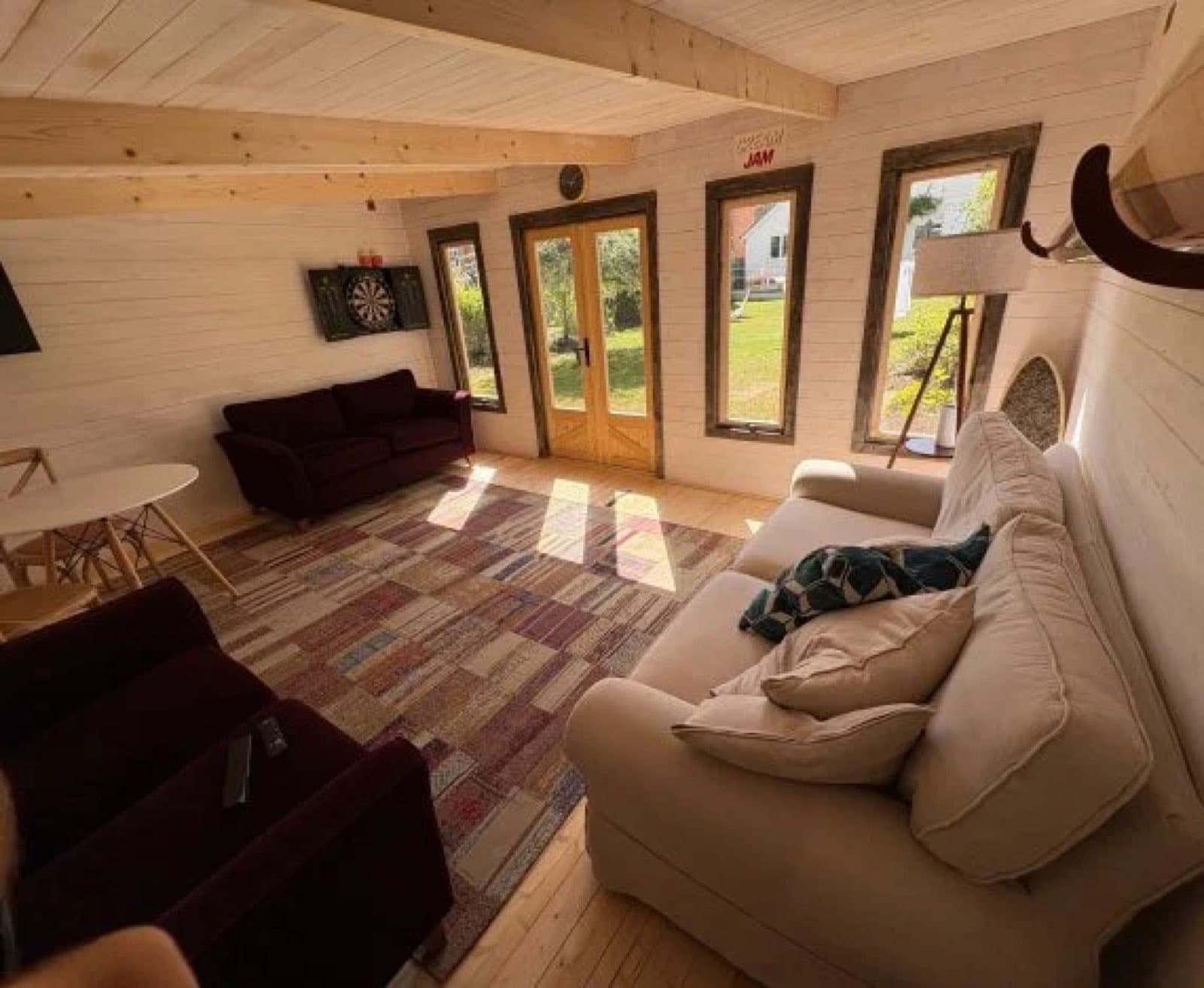 Inside a white painted log cabin lounge with two sofas, a dartboard, a rug and glazed doors onto the lawn