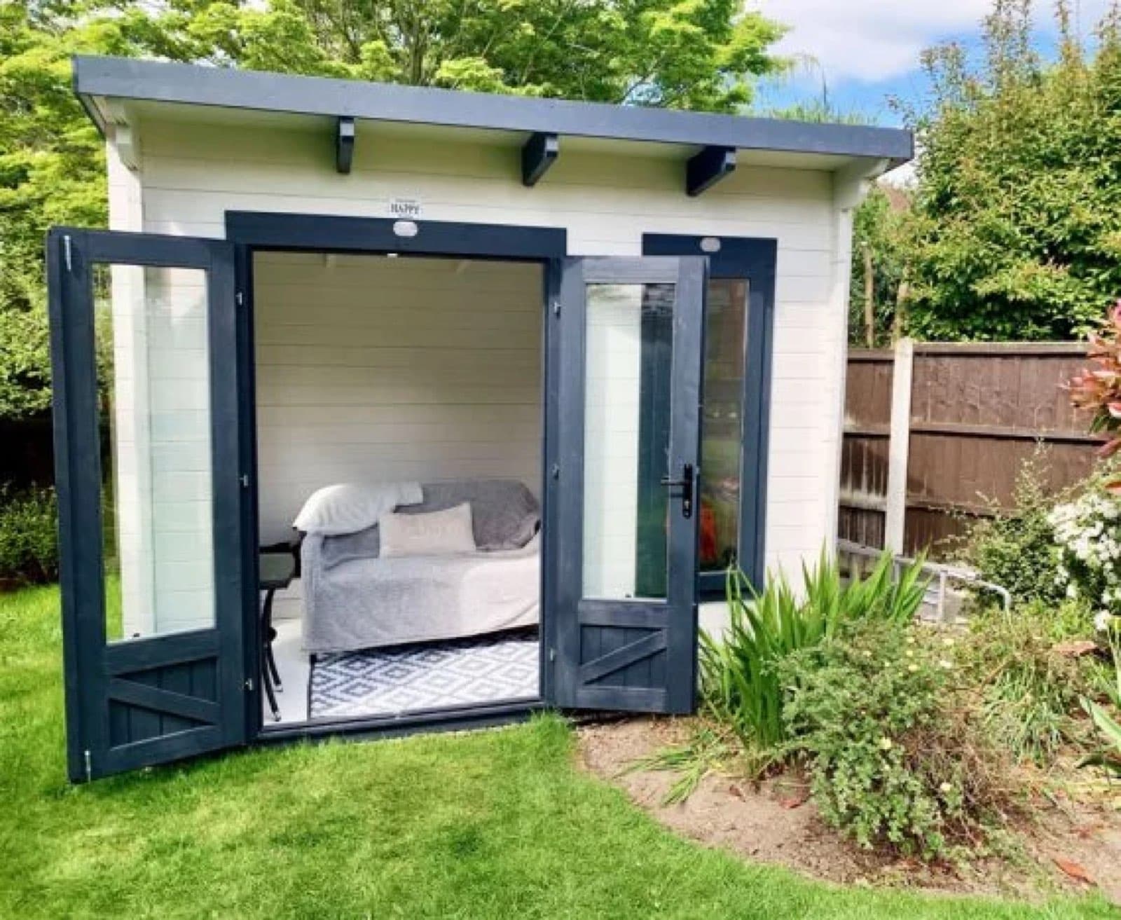 Doors open onto a day bed, for a spare room in the garden. Cream painted pent log cabin with navy doors open to show a day bed inside, on a lawn beside a border
