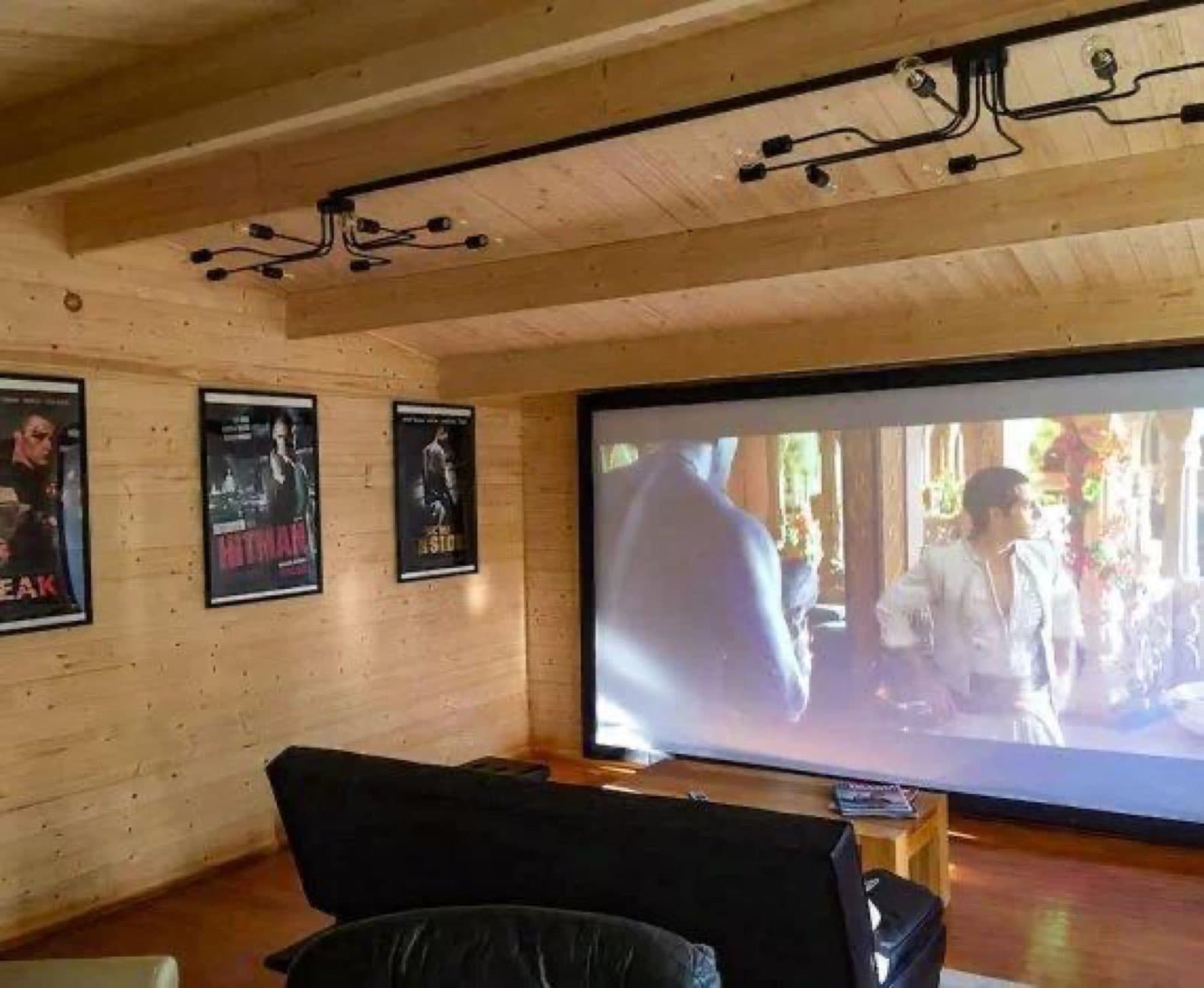 Log cabin cinema room with a wall-sized screen. Inside a log cabin cinema room with a large projector screen, framed film posters and pine-lined walls
