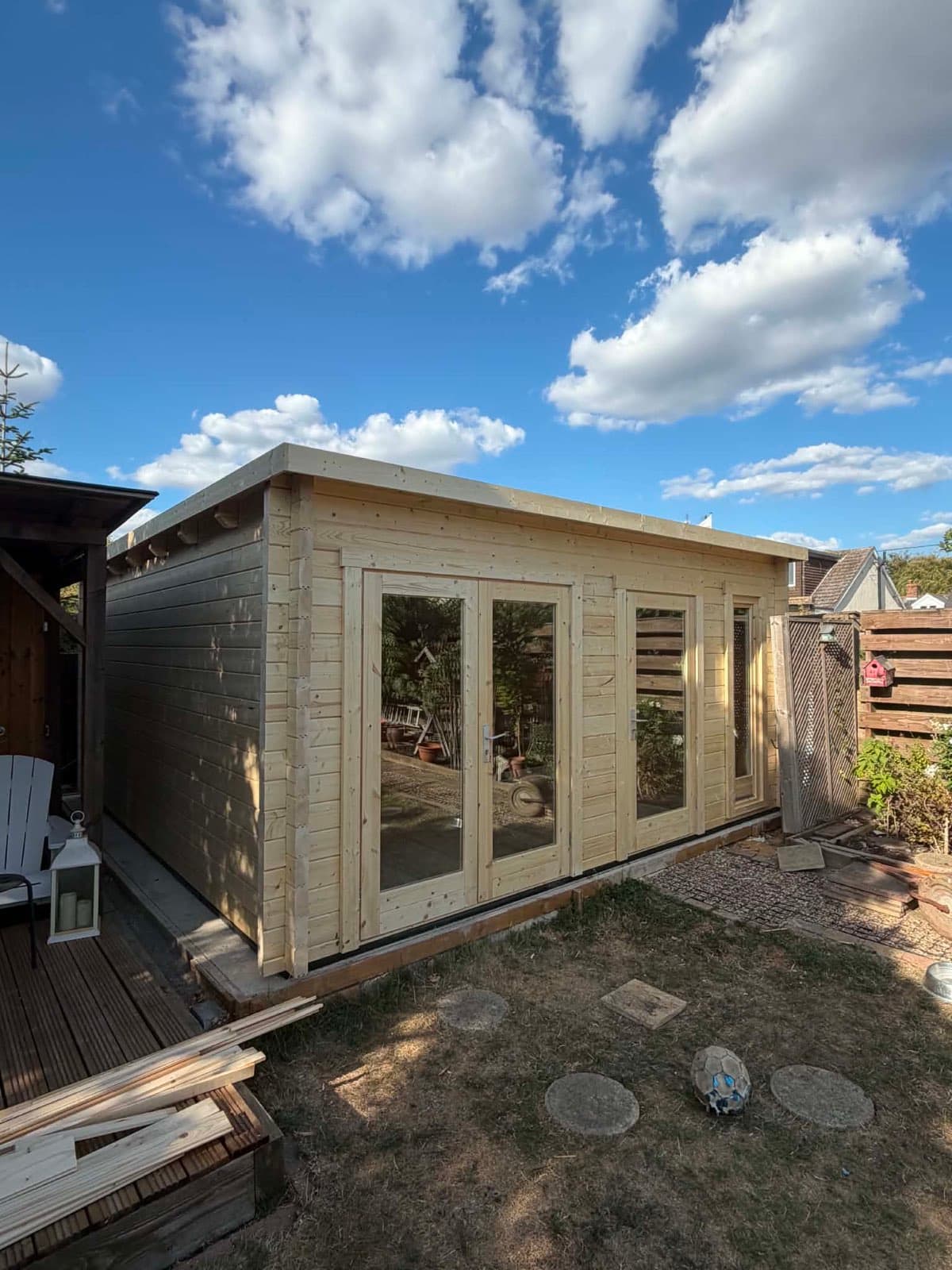 Pent-roof cabin, finished and ready to use. Untreated pent-roof log cabin with glazed double doors and side windows, beside timber decking under a blue sky