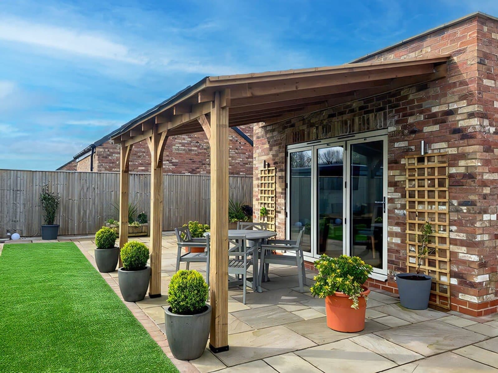 Lean-to oak pergola shading a sandstone patio. Oak lean-to pergola fixed to a brick house over a sandstone patio, with clipped topiary in pots and bifold doors behind