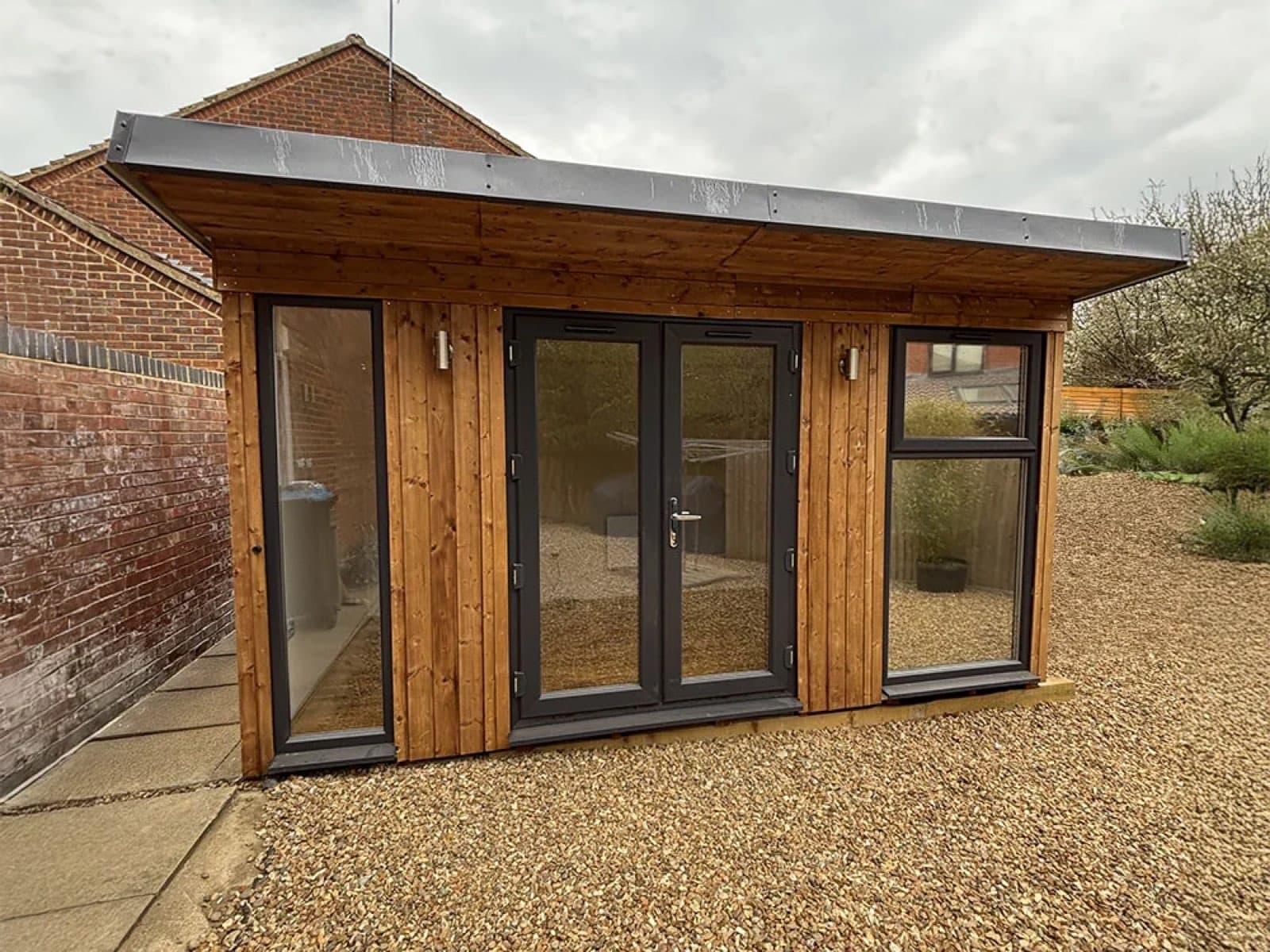 Timber-clad garden room with aluminium bifold doors. Timber-clad garden room with a grey zinc-effect roof and anthracite aluminium bifold doors, on a gravel base beside a brick wall