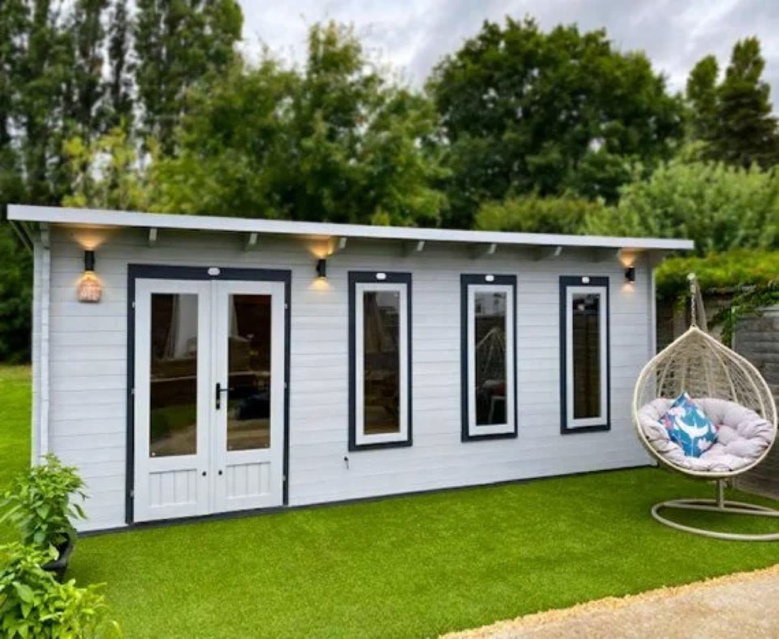 Long white cabin with a hanging chair alongside. White and grey painted pent log cabin with a run of windows and glazed doors, with a hanging egg chair on artificial lawn alongside